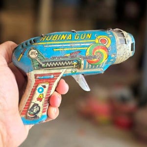 Pode incluir: Pistola de brinquedo vintage com o texto "RUBINA GUN" em amarelo. A pistola é predominantemente azul com detalhes em vermelho, branco e verde. Possui um gatilho e um design espiral colorido. O brinquedo é segurado em uma mão.