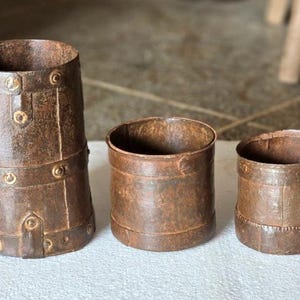 May include: Three antique, cylindrical metal containers with a rusted brown finish. The tallest has decorative metal bands and rivets. The other two are smaller and simpler in design. They are likely vintage or rustic decor.