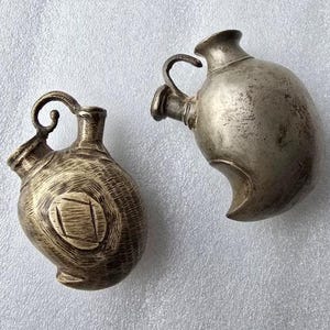 May include: Two antique metal pitchers with handles. One pitcher has a decorative swirl on the handle and a carved design on the body. The other pitcher has a simple handle and a curved base. Both pitchers have a tarnished, aged appearance.