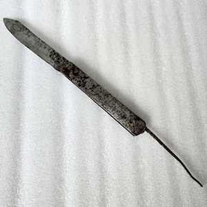 Könnte beinhalten: Ein antikes, langes, schlankes Metallwerkzeug mit spitzem Ende. Das Werkzeug hat eine flache, rechteckige Klinge mit leicht gebogener Spitze. Das Metall hat ein verwittertes, gealtertes Aussehen mit Gebrauchsspuren und Verfärbungen.