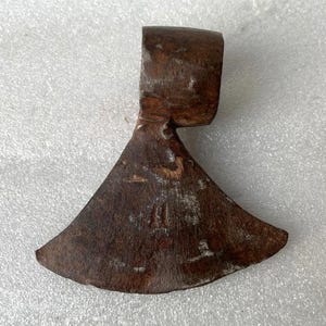 Peut inclure: Une tête de hache rouillée, avec une lame incurvée et un œil rectangulaire pour le manche. La tête de hache est de couleur marron foncé, avec des signes d'usure et de vieillissement, suggérant une utilisation pour couper du bois.