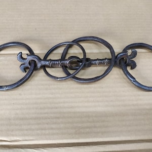 May include: A set of three interlocking metal rings with a key-like design. The rings are connected by a metal bar with a decorative pattern. The rings are made of dark metal and have a rustic appearance.