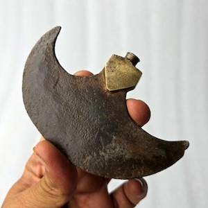May include: A crescent-shaped, antique metal blade with a brass handle. The blade has a dark, aged appearance, showing signs of wear and use. The brass handle is rectangular with a small, cylindrical top.