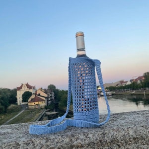 Könnte beinhalten: Ein hellblauer gehäkelter Weinflaschenhalter mit einem langen Riemen. Der Halter hält eine Weinflasche. Die Flasche steht auf einer Steinoberfläche. Der Hintergrund ist eine verschwommene Ansicht einer Stadtkulisse und eines Flusses.