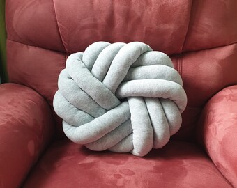 May include: A light gray knotted pillow on a red armchair. The pillow is made of soft fabric and has a unique knotted design.