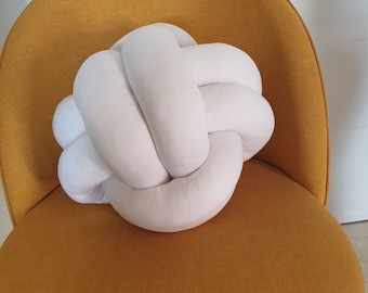 May include: A white knotted pillow sits on a mustard yellow chair. The pillow is made of soft, plush fabric and has a unique knotted design.