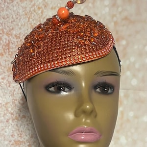May include: An orange-toned, embellished beret-style hat. The hat is covered in small, sparkling crystals and features a decorative beaded accent on top. The hat is displayed on a mannequin head with makeup.