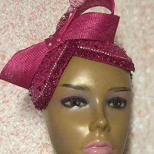 May include: A magenta-colored headpiece with a decorative bow and beaded accents. The headpiece features a textured fabric and is adorned with small, sparkling beads along the edges. A decorative element with pink and white stripes adds a touch of elegance.
