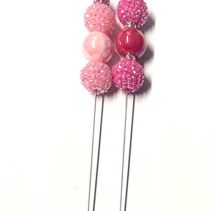 Könnte beinhalten: Zwei dekorative Hutnadeln mit rosa und irisierenden Perlen. Jede Nadel hat eine Reihe von Perlen in verschiedenen Rosatönen, darunter glitzernde, irisierende und unifarbene Farben. Die Nadeln sind aus silberfarbenem Metall.