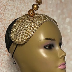 Puede incluir: Diadema dorada con un diseño decorativo de pequeñas cuentas y un acento de bola dorada. La diadema se lleva en una cabeza de maniquí.