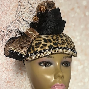 May include: A fascinator hat featuring a leopard print base, adorned with a black and bronze bow, black netting, and rows of small black and clear rhinestones. The hat is displayed on a mannequin head with brown eyes and pink lips.