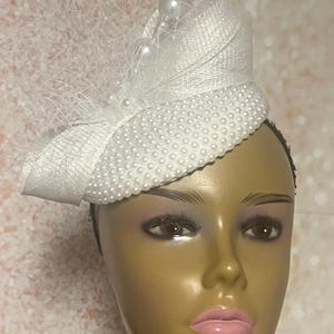 Peut inclure: Un chapeau blanc orné d'un nœud et de perles. Le chapeau est doté d'un voile en résille et est orné de petites perles sur le bord et d'un groupe de perles plus grosses. Le chapeau est conçu pour être porté sur la tête.