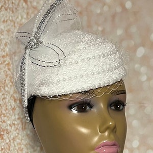 Puede incluir: Un sombrero blanco con velo y adornos de perlas. El sombrero tiene un ribete plateado y un lazo en un lado.