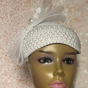 Può includere: Un cappello bianco con un design di perle e un fiocco di tulle bianco. Il cappello è realizzato in un materiale morbido e leggero, perfetto per un'occasione speciale.