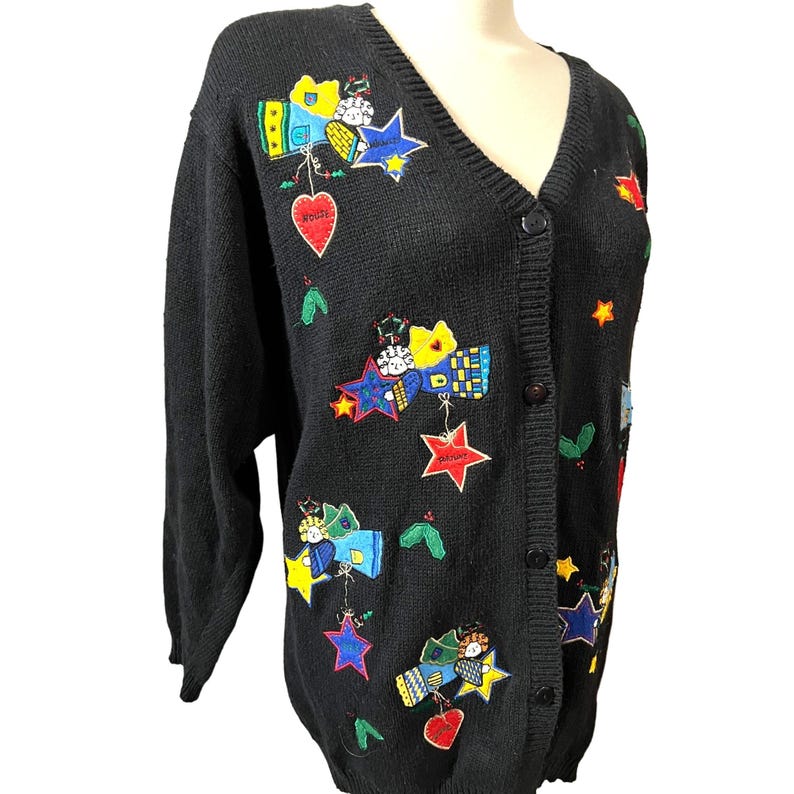 K&ouml;nnte beinhalten: Schwarzer Strickcardigan mit V-Ausschnitt und Knopfleiste. Der Cardigan ist mit bunten gestickten Engeln, Sternen und Herzen verziert. Der Text "Joy" und "Peace" ist auf die Herzen gestickt.