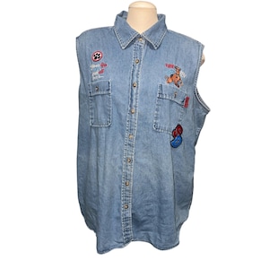 May include: Light blue denim sleeveless shirt with button-up front, two chest pockets, and embroidered patches. Patches include a paw print, a cartoon dog, and other colorful designs. The shirt has a collared neckline.
