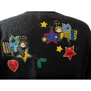 K&ouml;nnte beinhalten: Schwarzer Strickpullover mit bunten gestickten Engeln, Sternen und Herzen. Die Herzen sind mit den Worten "Love" und "Fame" bestickt.