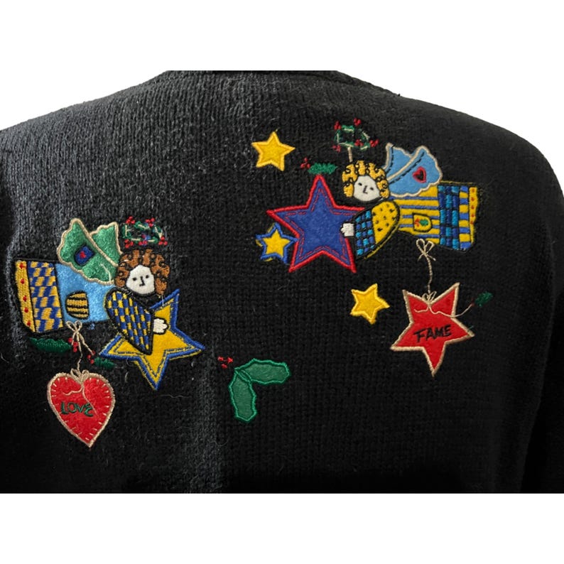 K&ouml;nnte beinhalten: Schwarzer Strickpullover mit bunten gestickten Engeln, Sternen und Herzen. Die Herzen sind mit den Worten "Love" und "Fame" bestickt.