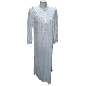 Puede incluir: Un camisón blanco con bordado floral azul y finas rayas azules. El camisón tiene un frente abotonado y un cuello con volantes.