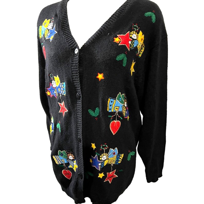 K&ouml;nnte beinhalten: Schwarzer Cardigan-Pullover mit Weihnachtsmotiv. Der Pullover zeigt bunte gestickte Engel, Sterne und Stechpalmenbl&auml;tter.