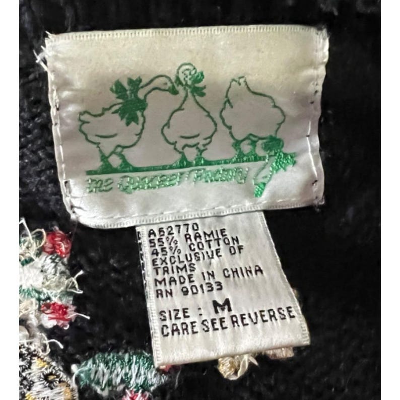 K&ouml;nnte beinhalten: Ein schwarz-wei&szlig;es Etikett mit dem Text "The Rocket Factory" und einer Grafik von drei G&auml;nsen. Das Etikett enth&auml;lt auch den Text "A52770 55% Ramie 45% Baumwolle Exklusiv von Bes&auml;tzen Hergestellt in China RN 90133 Gr&ouml;&szlig;e: M Pflegehinweise siehe R&uuml;ckseite".