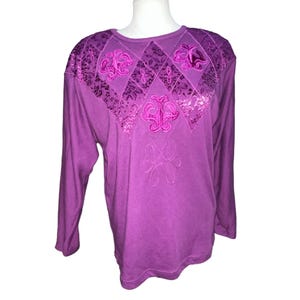 Puede incluir: Una camisa de manga larga de color morado con un diseño floral bordado en la parte delantera. El bordado es de un tono morado más oscuro.