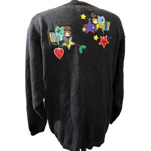 K&ouml;nnte beinhalten: Schwarzer Strickpullover mit einem weihnachtlichen Design mit gestickten Sternen, Herzen und Engeln. Das Design ist in Rot-, Gr&uuml;n-, Gelb-, Blau- und Wei&szlig;t&ouml;nen gehalten.