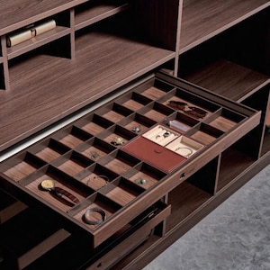 May include: A dark wood jewelry drawer organizer with multiple square compartments, featuring a brown leather-covered insert, a wristwatch with a brown leather strap, a silver bracelet, and various small rings and accessories arranged inside.