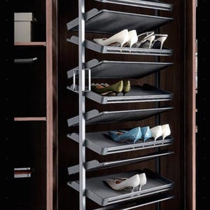 Op de afbeelding: Een hoog, verticaal uittrekbaar schoenenrek met een zilverkleurig metalen frame en meerdere schuine planken. De planken bevatten diverse paren schoenen met hakken in wit, olijfgroen, blauw en beige, georganiseerd in een donkerhouten kast.