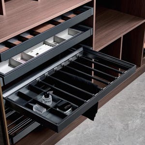 Puede incluir: Un sistema de cajones extraíbles de color gris oscuro para armario con múltiples compartimentos. El cajón superior cuenta con divisores claros para joyas, mientras que el inferior incluye un riel para colgar prendas y secciones para frascos.