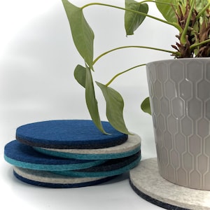 May include: A stack of round felt coasters in shades of blue, gray, and white. A potted plant with green leaves is in the background. The coasters are stacked on top of each other, with a single coaster in the foreground.