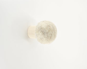 May include: A light-colored, round felt ball attached to a small, light-colored wooden base. The felt ball has a textured, woolly appearance. The wooden base is a flat, circular shape. The object is set against a white background.