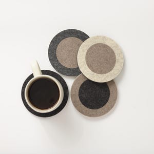 May include: A set of four circular felt coasters in shades of charcoal, light gray, and beige, arranged in a cluster. One coaster supports a white ceramic mug filled with dark coffee. The coasters feature a two-tone design with a solid center circle.
