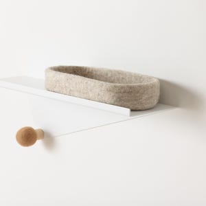 May include: A white shelf with a light brown felt basket and a wooden knob. The shelf is angled and mounted to a white wall.