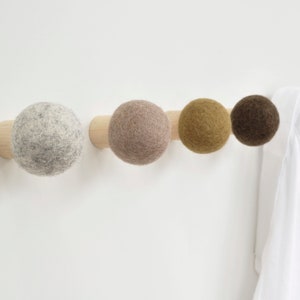 May include: A wooden wall-mounted coat rack with four felt ball hooks in shades of gray, mauve, gold, and brown. A white shirt hangs on the rightmost hook. The rack is mounted on a white wall.