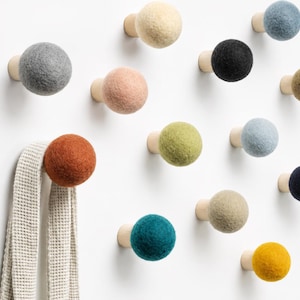 May include: A collection of wall-mounted hooks featuring spherical felted wool heads in various colors including gray, cream, pink, black, blue, olive, rust, teal, and yellow, each attached to a light-colored wooden base, with a beige waffle-knit towel.