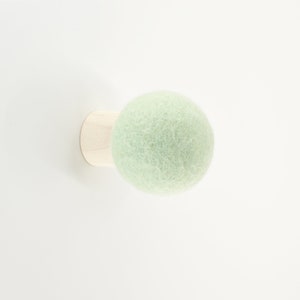 May include: A light green felt ball attached to a small, round, natural wood base. The felt ball is a soft, fuzzy texture. The wood base is a light tan color. The object is set against a plain white background.