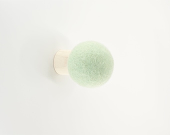 May include: A light green felt ball attached to a small, round, natural wood base. The felt ball is a soft, fuzzy texture. The wood base is a light tan color. The object is set against a plain white background.