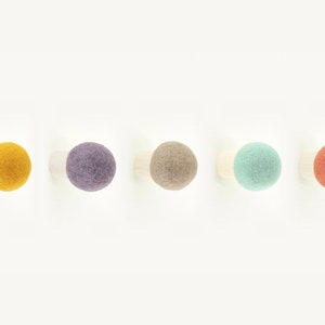 May include: Five felt ball wall hooks in different colors: yellow, purple, beige, light blue, and orange. The hooks are mounted on a white wall.