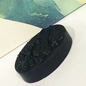 May include: A black oval-shaped bar of soap with a detailed relief design of bees and flowers. The soap has a matte finish and is set against a white and watercolor background.