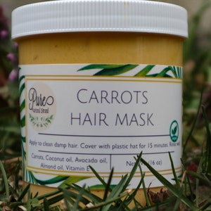 May include: A jar of carrot hair mask with a white lid and a label that reads "Carrots Hair Mask." The label also lists ingredients like carrot, coconut oil, and avocado oil. The jar is sitting in grass.
