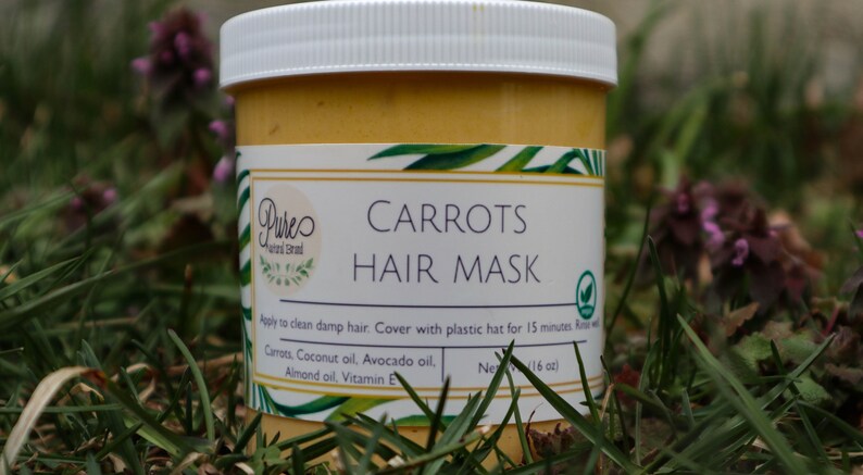 May include: A jar of carrot hair mask with a white lid and a label that reads "Carrots Hair Mask." The label also lists ingredients like carrot, coconut oil, and avocado oil. The jar is sitting in grass.