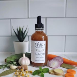 May include: A clear glass bottle with a black dropper, filled with a brown liquid, labeled "Pure Natural Brand Pre-Poo Dropper." The image includes fresh herbs, vegetables, and a small potted plant, suggesting natural ingredients.