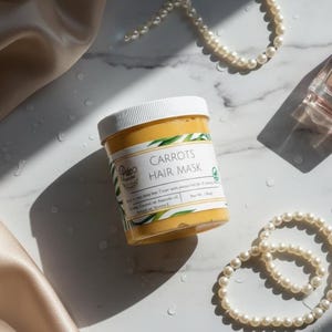 May include: A jar of "Carrots Hair Mask" with a white lid, set on a marble surface. The jar is surrounded by a pearl necklace, a bottle of perfume, and a satin cloth. The hair mask has a yellow-orange color.