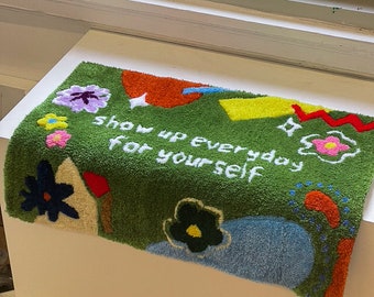 May include: Green rug with colorful abstract shapes and flowers. The rug has the text "Show up everyday for yourself" embroidered on it.