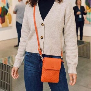 Può includere: Un cappello fedora arancione brillante, un cardigan beige con scollo a V e bottoni, un dolcevita nero, jeans blu scuro e una piccola borsa a tracolla arancione con trama intrecciata e chiusura a patta con bottone.