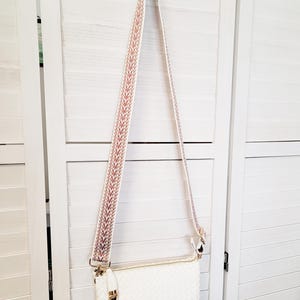 Pu&ograve; includere: Una borsa a tracolla rettangolare bianca con trama intrecciata e chiusura a zip color oro. La borsa presenta una tracolla lunga e regolabile con un motivo geometrico ripetuto nei toni dell'arancione, bianco e grigio, fissata con finiture dorate.