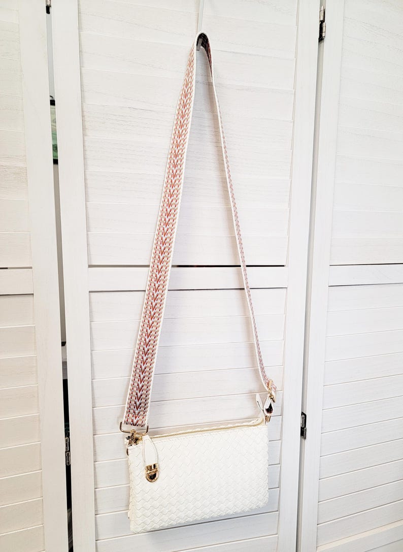 Pu&ograve; includere: Una borsa a tracolla rettangolare bianca con trama intrecciata e chiusura a zip color oro. La borsa presenta una tracolla lunga e regolabile con un motivo geometrico ripetuto nei toni dell'arancione, bianco e grigio, fissata con finiture dorate.