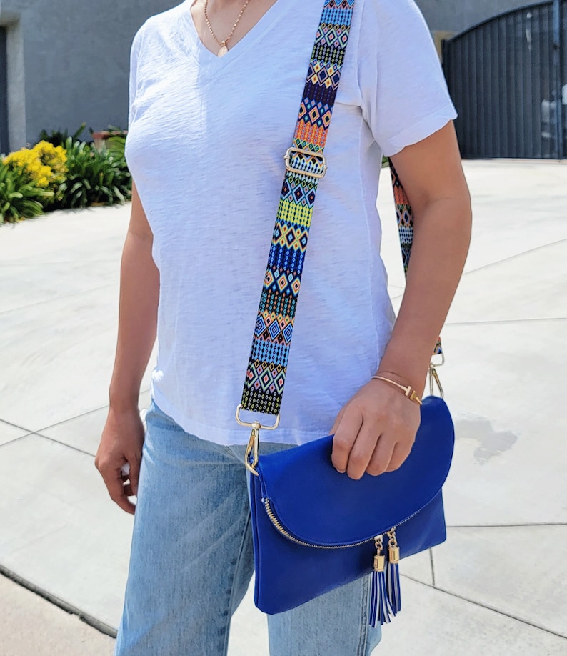 Pu&ograve; includere: Una borsa a tracolla blu vivace con patta, dotata di una cerniera color oro con due nappe abbinate a righe blu e bianche. La borsa include un'ampia tracolla regolabile decorata con un colorato e intricato motivo geometrico a rombi.