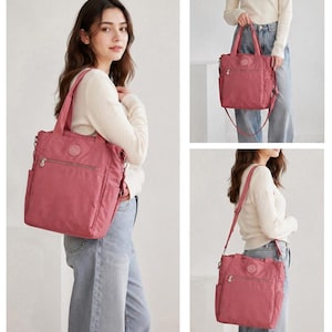 May include: A dusty rose nylon tote bag featuring a front zippered pocket, two top handles, and a detachable shoulder strap. The bag includes side pockets and a circular logo patch on the front, designed for versatile carrying options.
